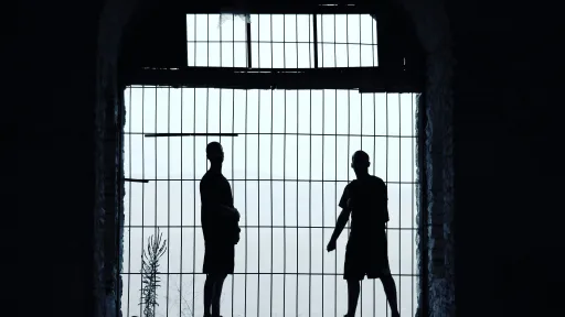 Two silhouetted human figures stand behind the bars of a prison gate